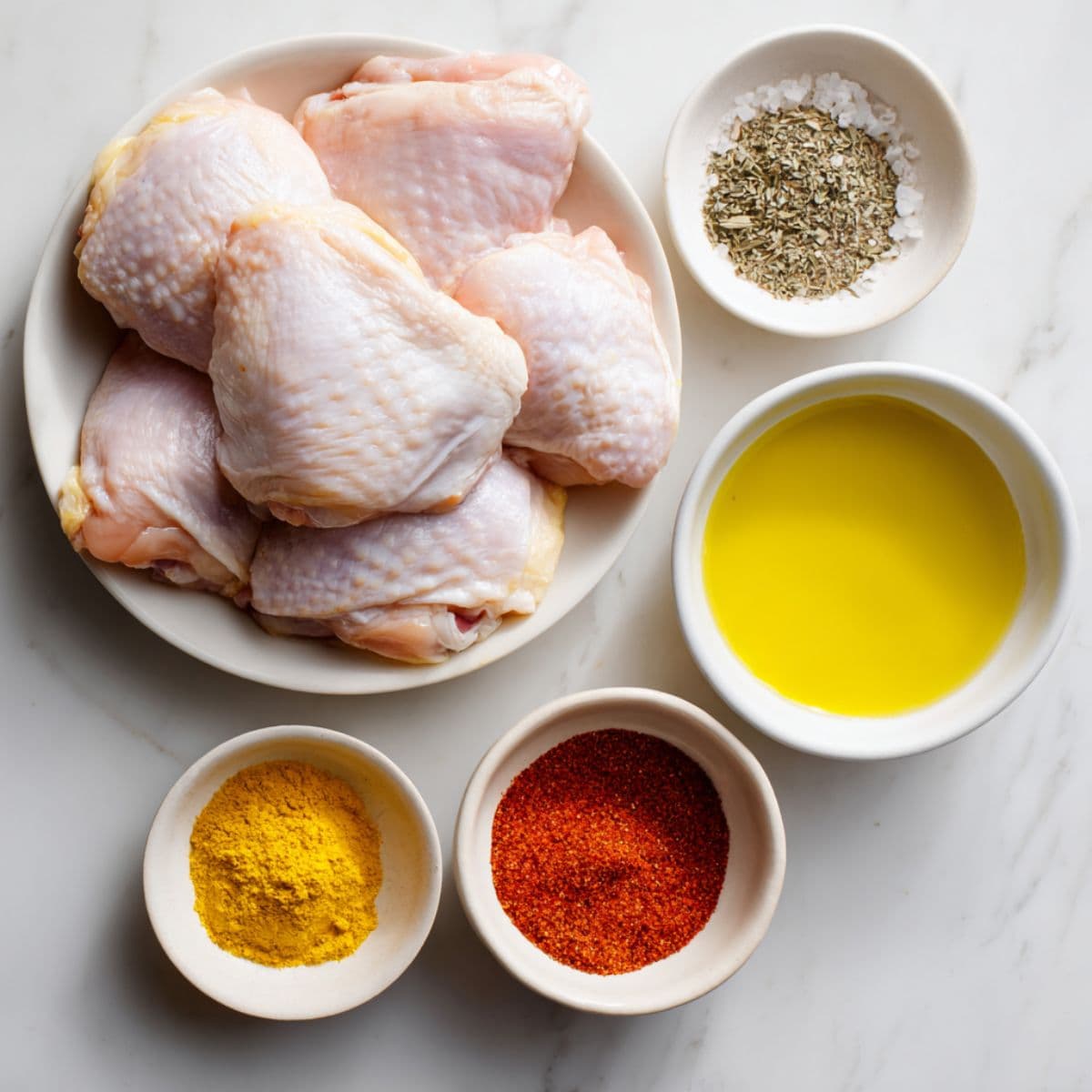 raw bone-in, skin-on chicken thighs arranged on white marble with small bowls of olive oil, kosher salt, black pepper, garlic powder, and paprika