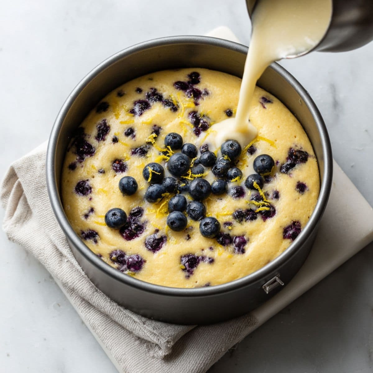 lemon blueberry cake batter poured into a prepared baking pan with visible blueberries, ready to bake on white marble