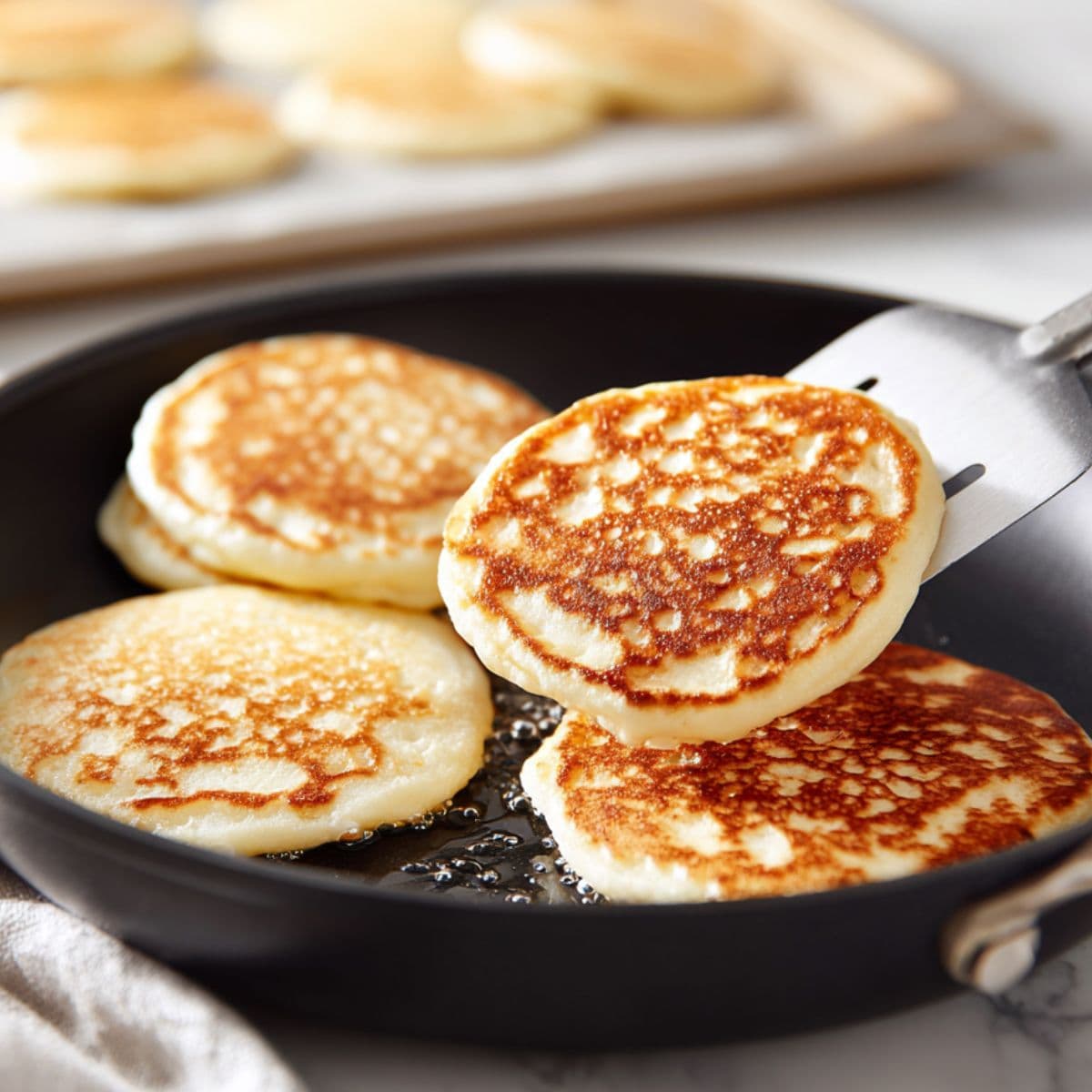 golden brown protein pancakes cooking in non-stick skillet with spatula gently lifting one pancake mid-flip showing bubbles on the surface