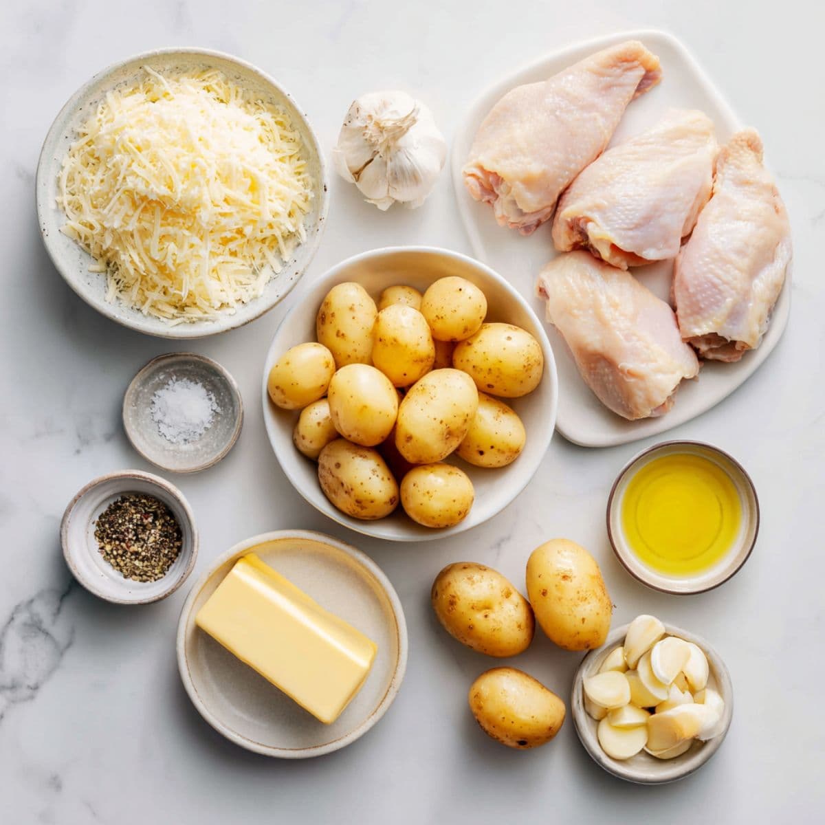 raw chicken breasts, baby potatoes, parmesan cheese, butter, olive oil, garlic cloves, salt, and pepper arranged neatly on white marble