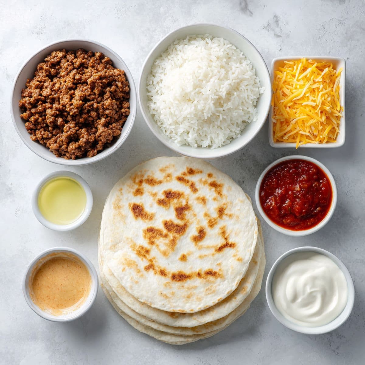 Flat lay of grilled cheese burrito ingredients on a light background ground beef, taco seasoning, flour tortillas, cooked rice, shredded cheddar and mozzarella, sour cream, and chipotle sauce