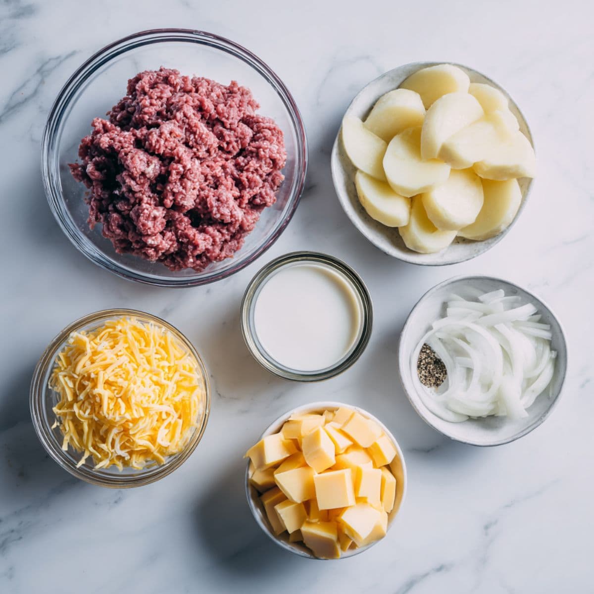 flat lay of ground beef, potatoes, onion, cheddar cheese, cream of mushroom soup, milk, and seasoning on white marble background