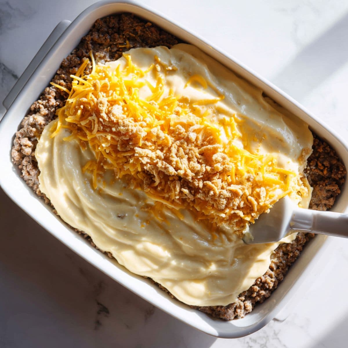 Overhead shot of a baking dish with the meatloaf layer topped with creamy mac and cheese, being spread to the edges and sprinkled with extra cheddar and panko before baking.